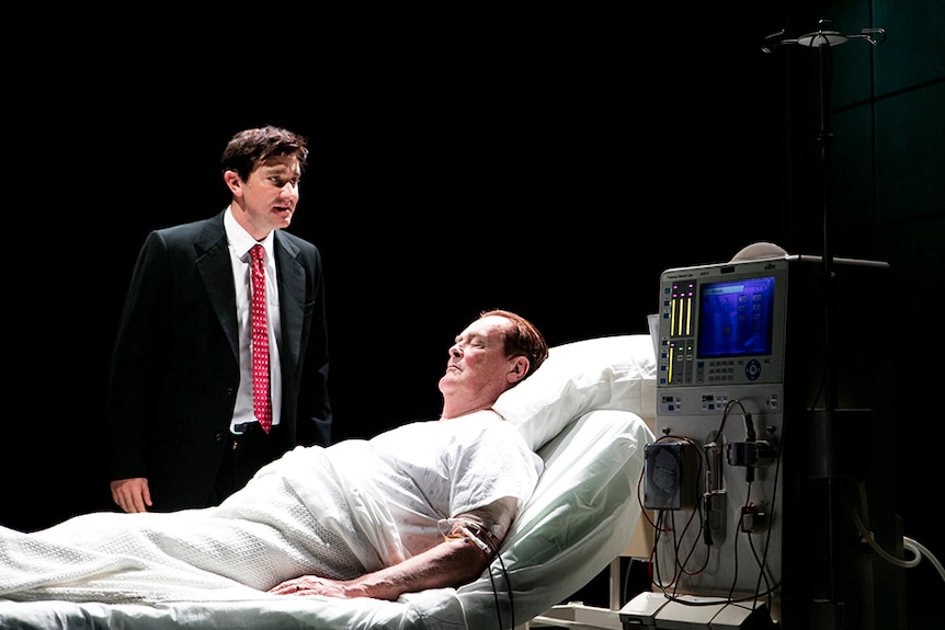 A distressed man wears dark suit with red tie and stands next to unconscious older man in hospital bed, connected to machine.
