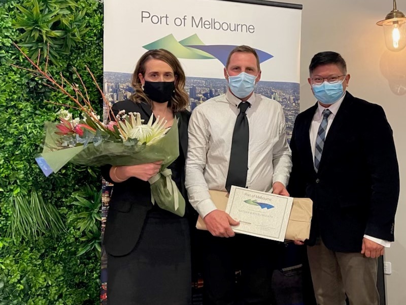 A woman holding a bunch of flowers and two men wearing face masks and holding an award.