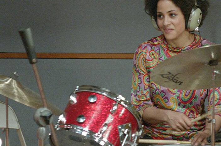 Colour still of a drummer Kimberly Thompson wearing large headphones, a brightly patterned top and playing the drums.