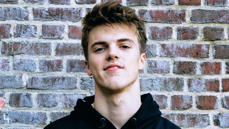 A young man wearing a black hoodie stands against a brick wall.