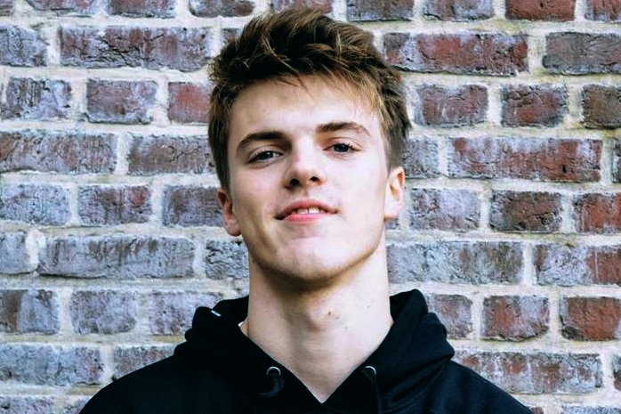 A young man wearing a black hoodie stands against a brick wall.