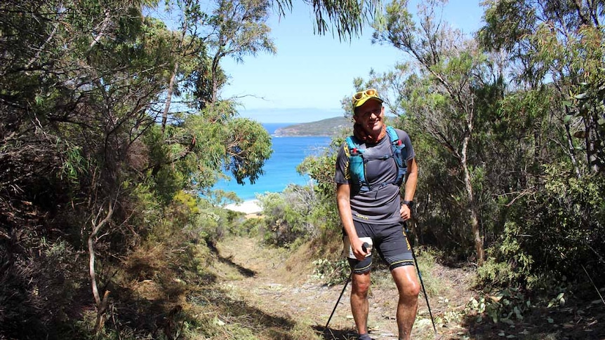 A man dressed in hiking gear stands on a trail overlooking the ocean.