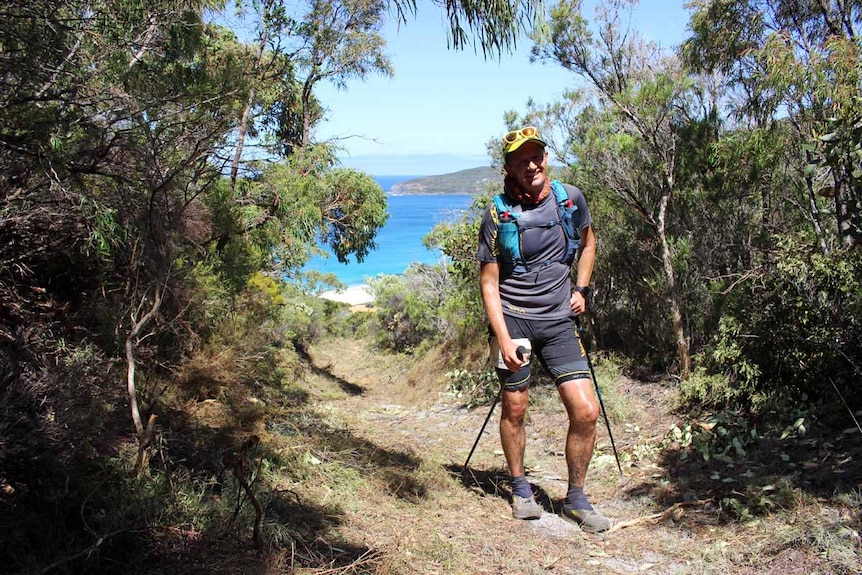 A man dressed in hiking gear stands on a trail overlooking the ocean.