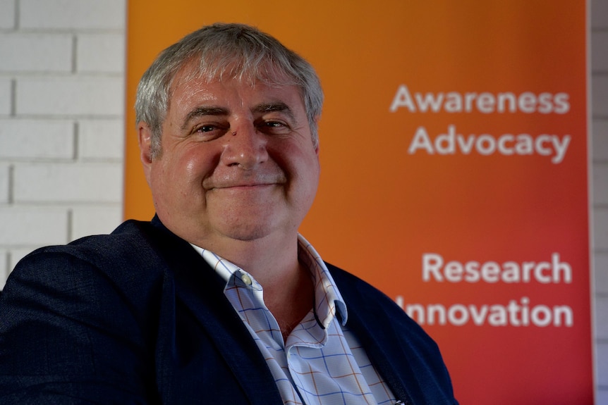 Man wearing suit smiles in front of orange banner with text saying awareness advocacy and research innovation.