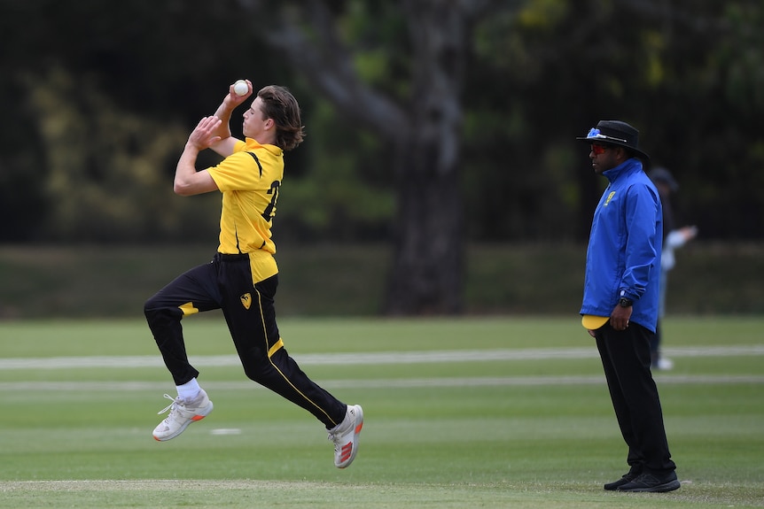 A cricketer wearing yellow and black runs in to bowl next to a umpire wearing a blue jacket