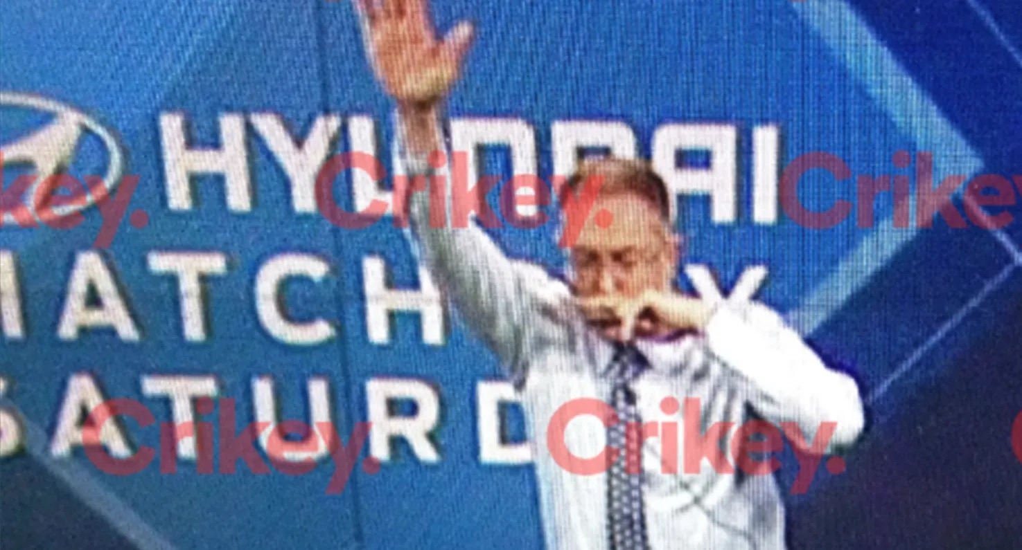 Foxtel CEO Patrick Delany with left hand in the air, right hand over top lip, blue and white signage behind. Crikey bug on image