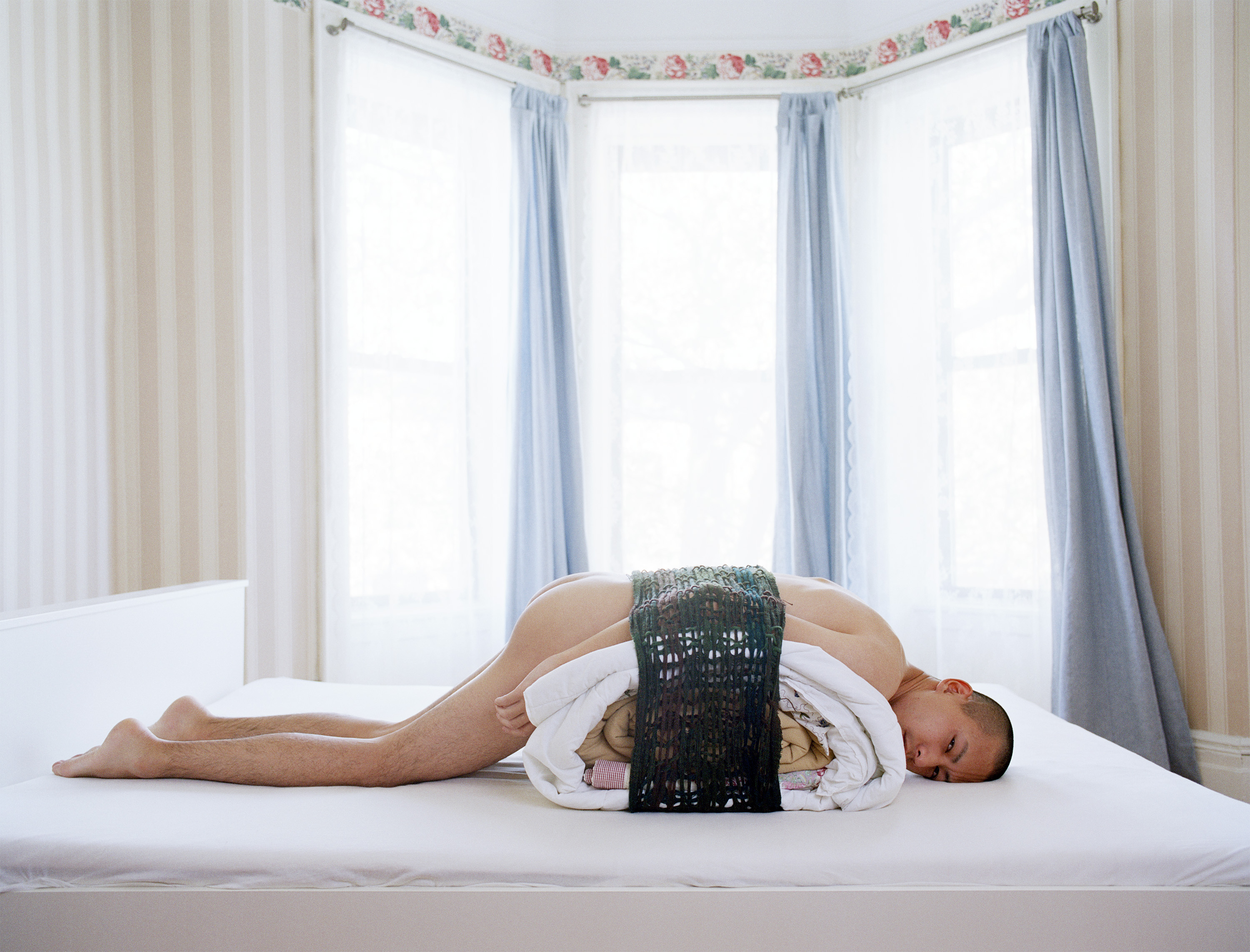 Moro, a Japanese man with a close-shaved hair cut in naked lying on a pile of sheets on a bed, styled as a sushi roll.