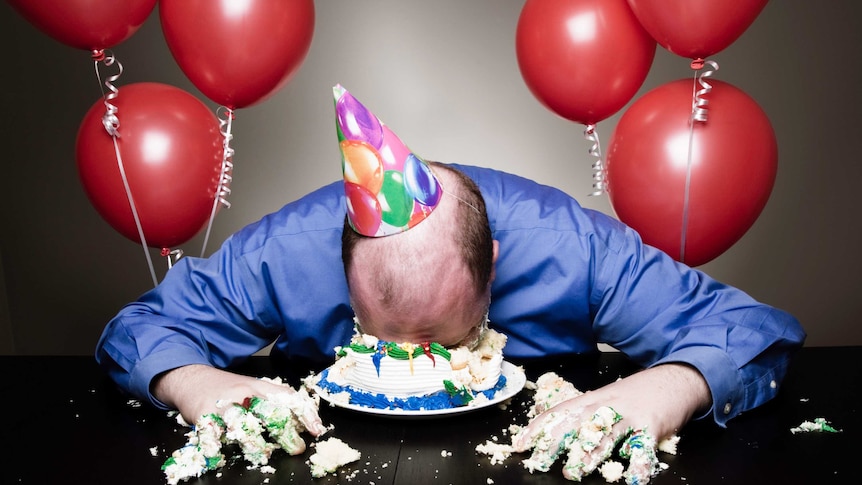 Balding man wearing party hat and blue shirt face first in a birthday cake surrounded by six red balloons
