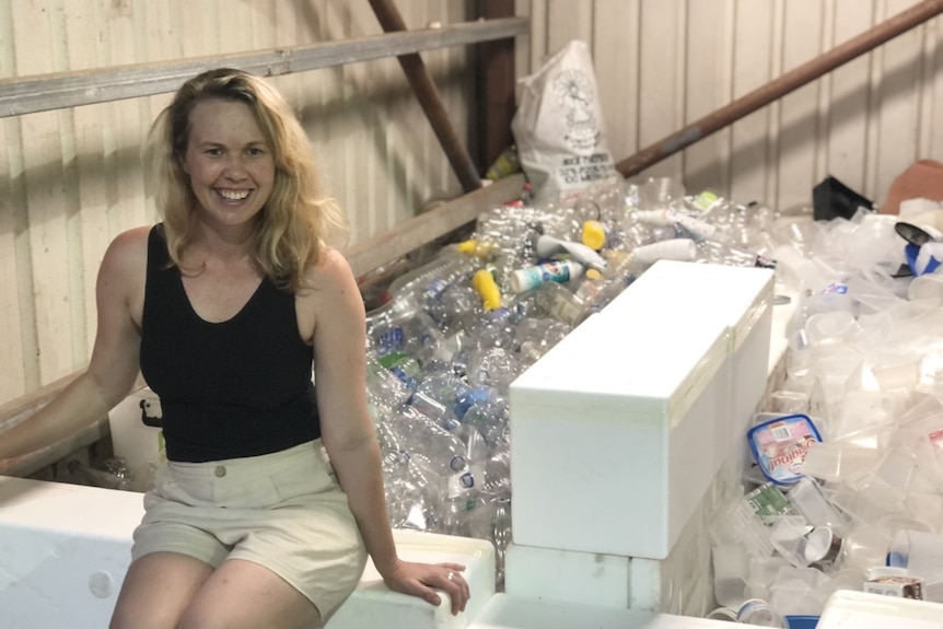Woman sits on Styrofoam box with masses of plastic bottles behind her. 