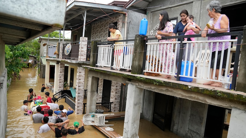 People on a first storey balcony watch others negotiate a flooded road, with some carrying belongings above the brown water.