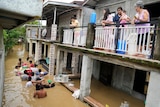 People on a first storey balcony watch others negotiate a flooded road, with some carrying belongings above the brown water.