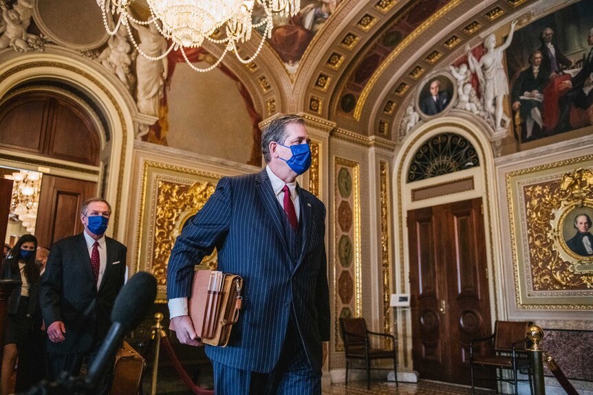 Castor wears a suit and mask and holds folders and a satchel as he walks through a grand hall past reporters