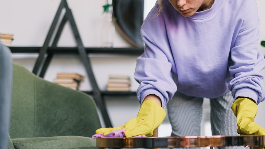 A woman in a purple jumper and yellow gloves wipes a small table