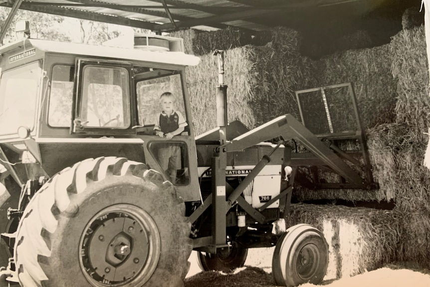 A young boy stands on a tractor under a shelter filled with hay bales. The image is black and white, the boy looks to camera