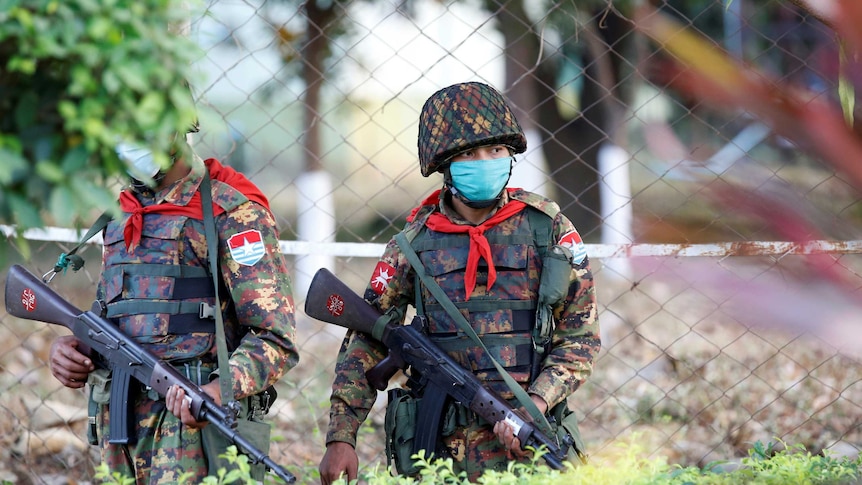 A soldier dressed in army uniform wears a mask and helmet and holds a gun in front of a fence.