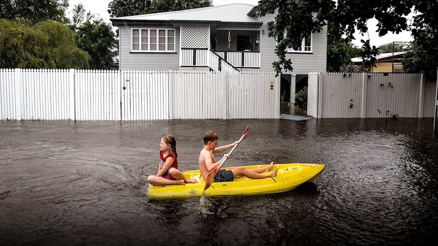 Two children paddle a yellow dinghy in floodwaters outside a suburban home