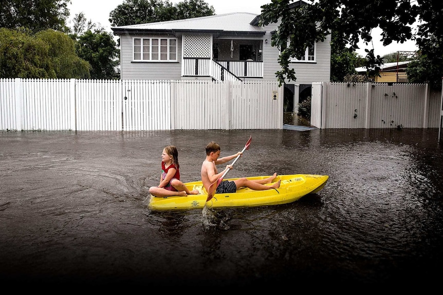 Two children paddle a yellow dinghy in floodwaters outside a suburban home