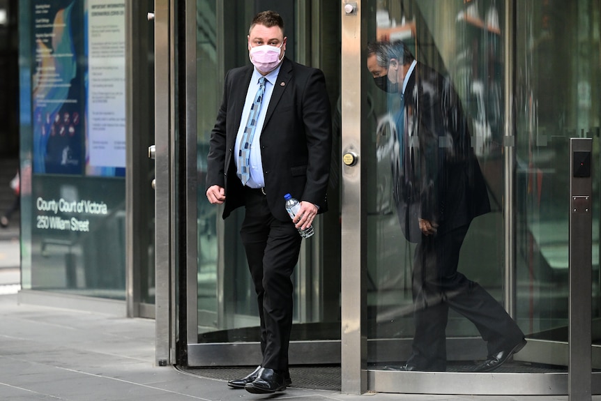 Brett Johnson wears a suit and a surgical face mask as he walks out the glass doors of a court.