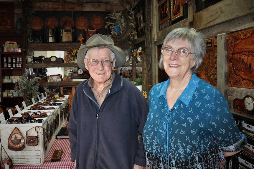 Elderly man in cowboy hat standing next to elderly lady on right in shop with shelves of leathers purses and bracelets.