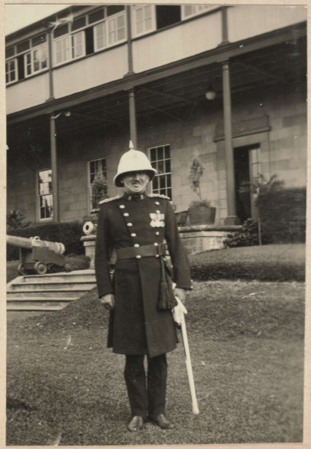 A male band member standing straight in position outside a barracks building in an old photo.