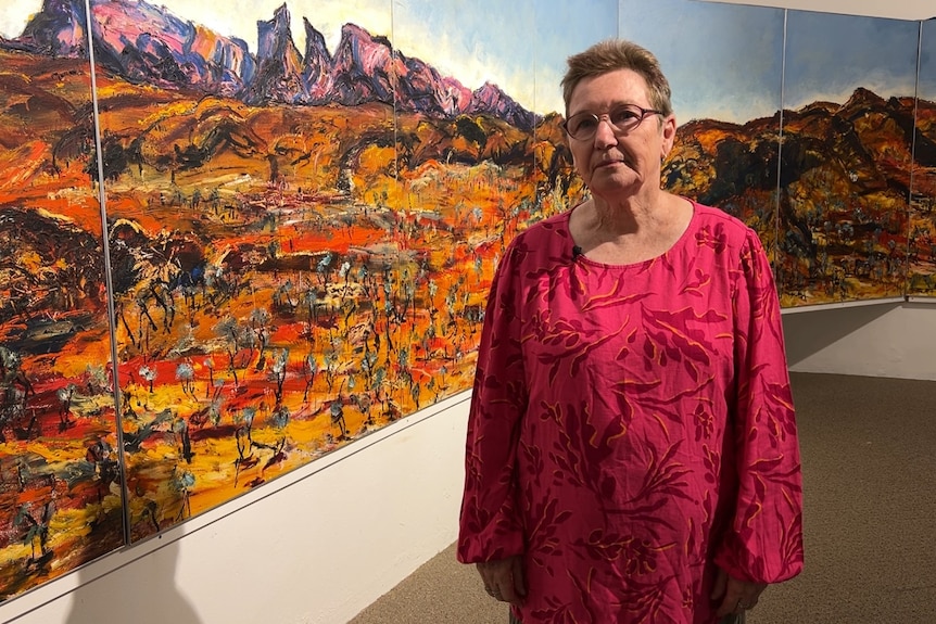 A woman in a pink shirt and glasses stands with a large mural of mountains behind her.