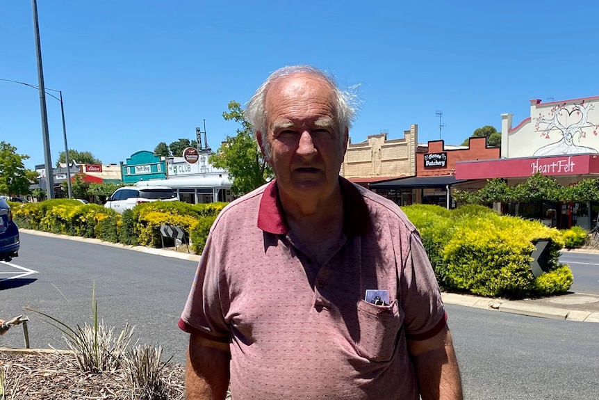 An old man stands on a regional main street with a nature strip and shops behind him.