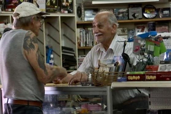 An older man smiles at a customer at the counter of a video store.