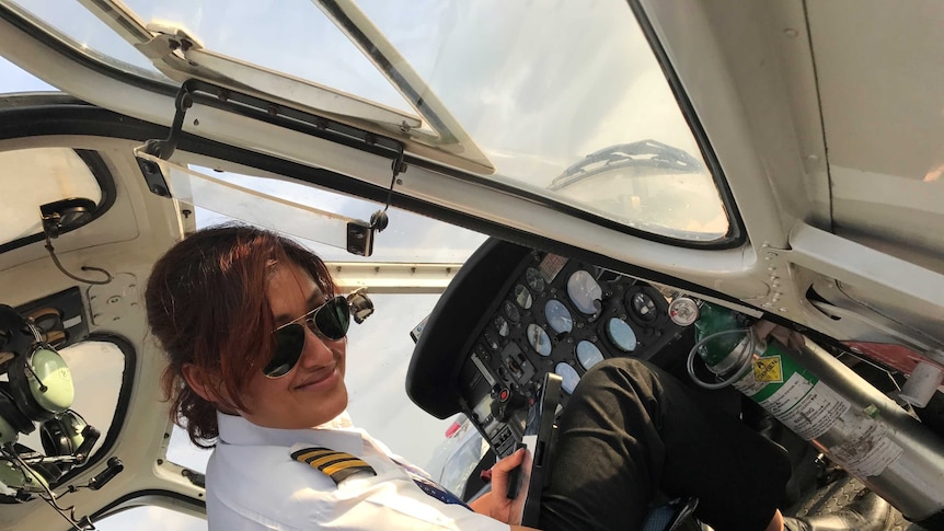 Woman wearing aviators and her captain stripes sits in the cockpit of a helicopter smiling.
