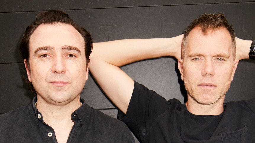 Two men wearing dark shirts standing before a dark wall. One has his hands behind his head.