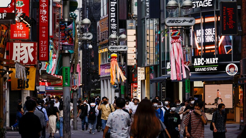 The very busy Shibuya district of Tokyo, crowds can be seen and everyone is outside and wearing masks.