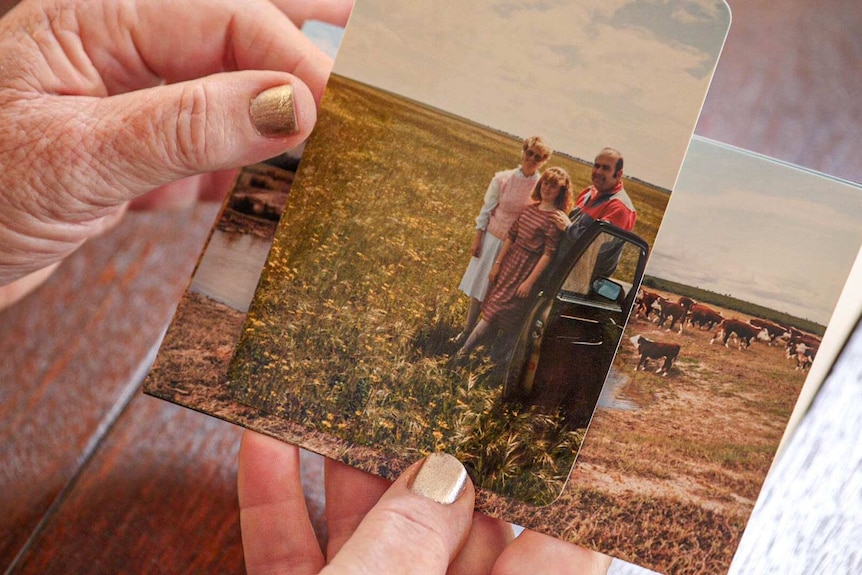 Hands with gold nail polish hold a stack of old photos, on top a photo of Heather Slade, her husband and daughter on their farm.