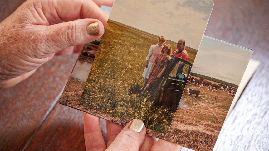 Hands with gold nail polish hold a stack of old photos, on top a photo of Heather Slade, her husband and daughter on their farm.