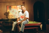 Musonga sitting on a pool table holding a pool cue with one of his illustrations behind him