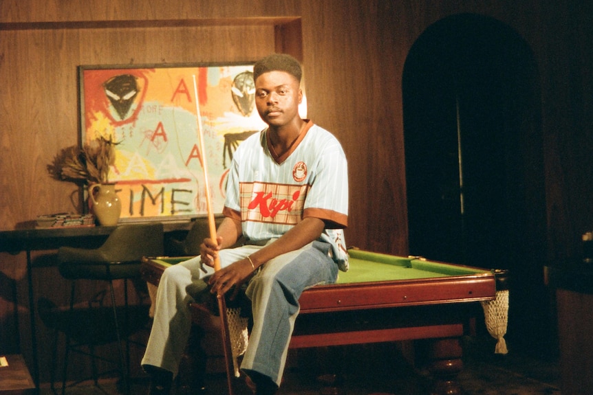 Musonga sitting on a pool table holding a pool cue with one of his illustrations behind him