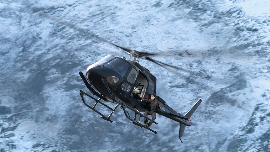 Colour still image of Tom Cruise hanging off the side of a helicopter in mid-air in 2018 film Mission Impossible: Fallout.