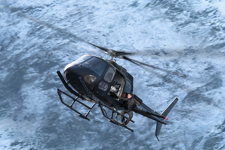Colour still image of Tom Cruise hanging off the side of a helicopter in mid-air in 2018 film Mission Impossible: Fallout.