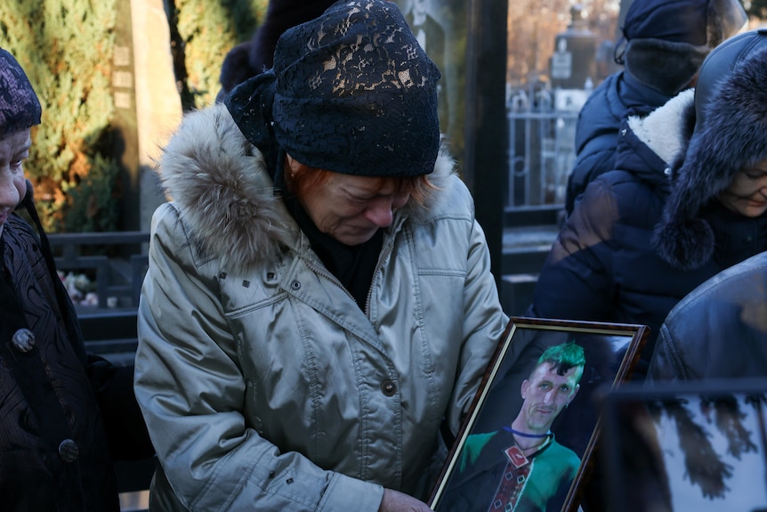 A woman wearing a beanie and gray puffer jacket lined with fur cries over a portrait of her son.