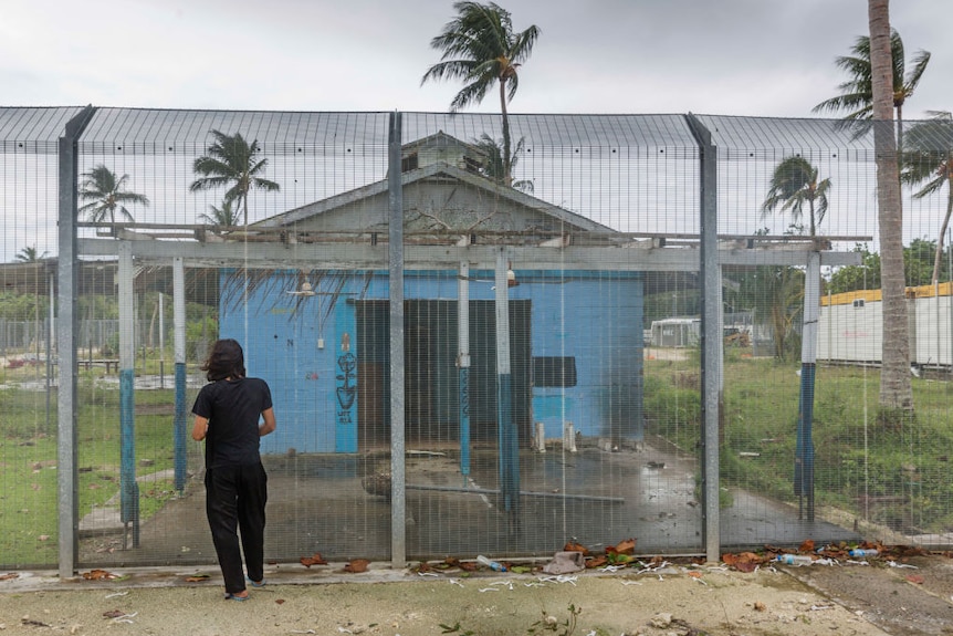 man with back turned faces towards worn down blue building guarded behind large security gate