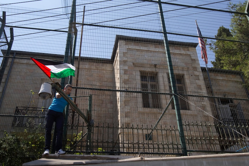 A man places a Palestinian flag on a fence surrounding a brick building with a US flag.