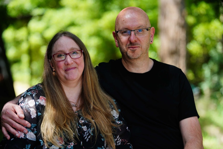 A couple stands with their arms around one another, looking towards the camera