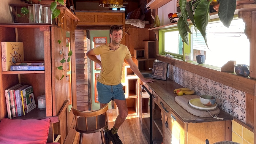 A man leans against a wooden benchtop in the middle of a tiny home. 