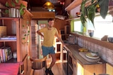 A man leans against a wooden benchtop in the middle of a tiny home. 