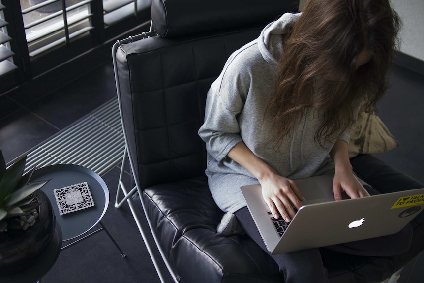 A woman whose hair covers her face sits on a leather chair working on an Apple laptop.