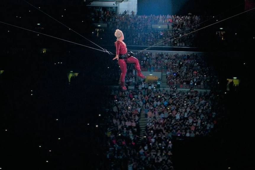 A blonde woman in a red leotard is wears a harness and is suspended above a concert crowd.