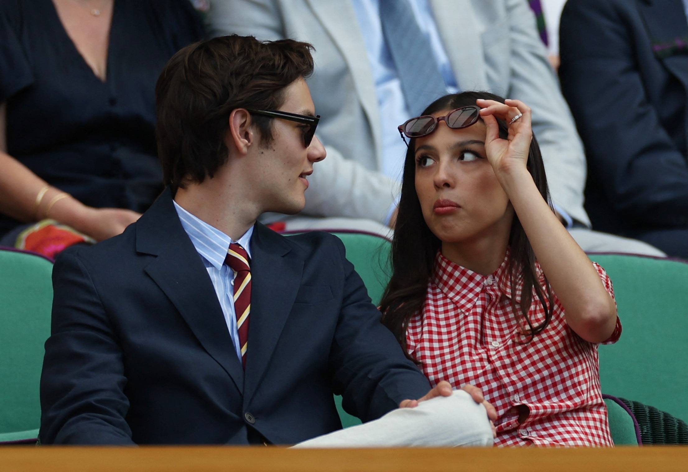 Olivia Rodrigo in a checked dress raises her sunglasses and pouts at her partner 