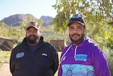 Two Indigenous men in jumpers smile at camera with a backdrop of mountain ranges