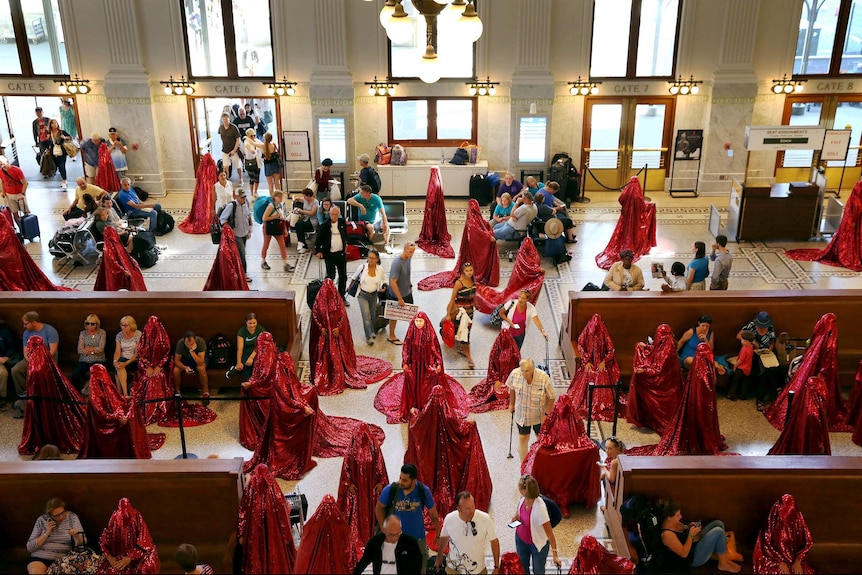 A group of people in sequinned red body coverings amongst a group of casually dressed passersby in a busy train station