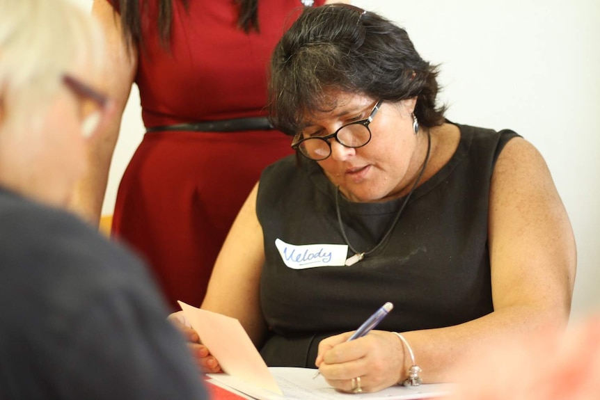 A middle-aged woman holds pen and paper and is concentrating on writing.