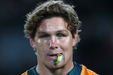 An Australian male rugby player in a Wallabies jersey walks with his mouthguard hanging out of his mouth.