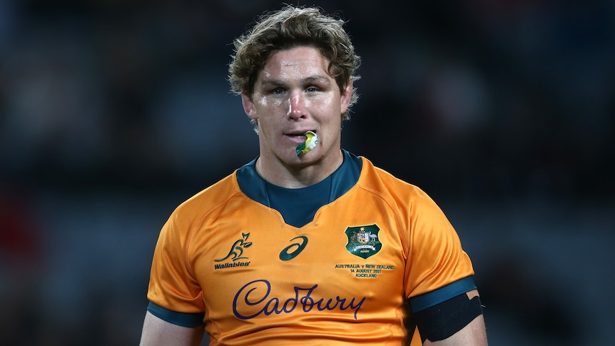 An Australian male rugby player in a Wallabies jersey walks with his mouthguard hanging out of his mouth.